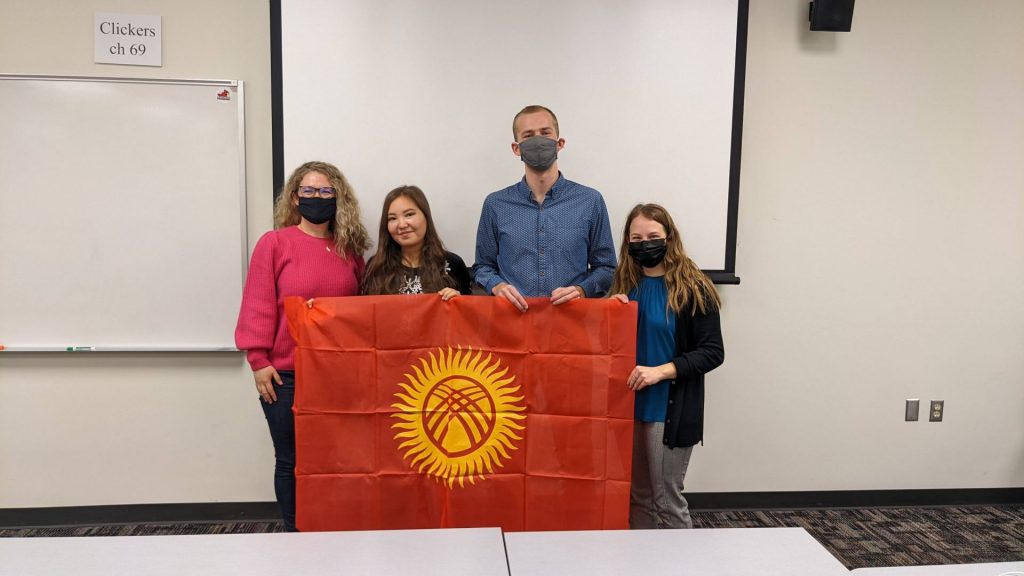 A faculty member and three students hold a flag at the front of a classroom.
